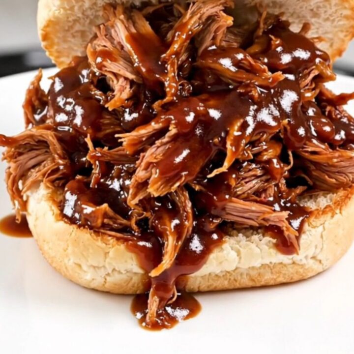 A close-up of glossy, shredded pulled pork piled high on a white bun on a plain round plate.