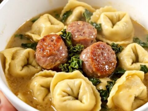 A steaming bowl of creamy sausage and kale tortellini held by a hand against a kitchen background.