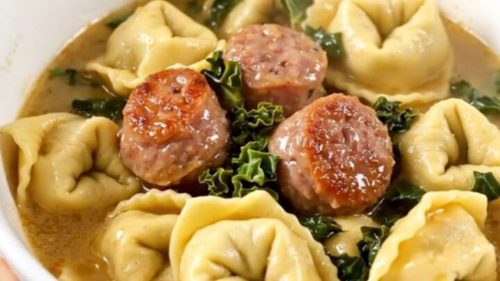 A steaming bowl of creamy sausage and kale tortellini held by a hand against a kitchen background.