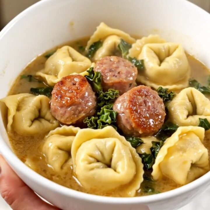 A steaming bowl of creamy sausage and kale tortellini held by a hand against a kitchen background.