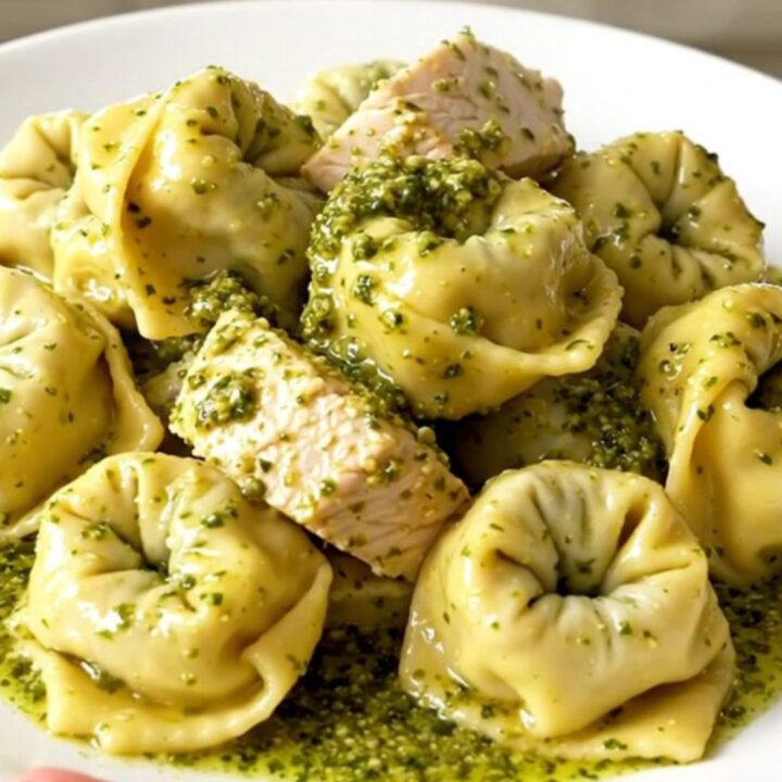 A close-up of a white plate filled with creamy pesto tortellini mixed with chicken and bacon.