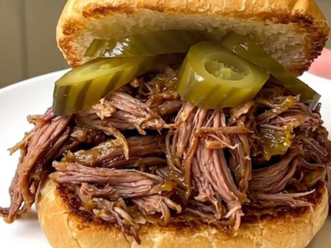 A close-up of a tender shredded beef sandwich on a toasted bun with pickle chips.