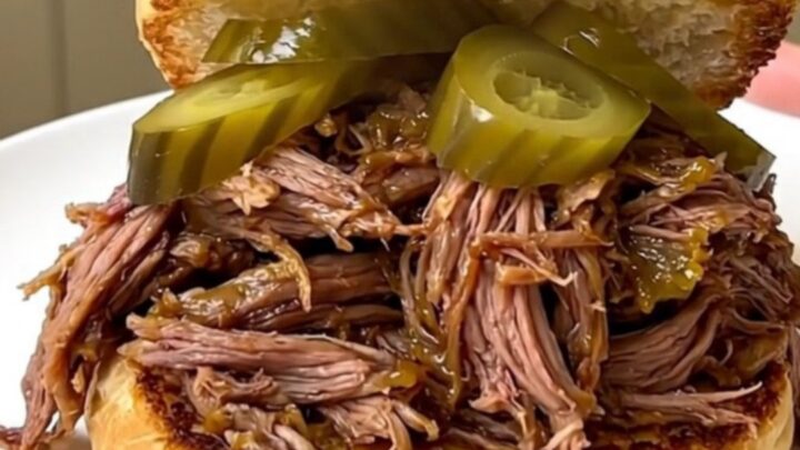 A close-up of a tender shredded beef sandwich on a toasted bun with pickle chips.