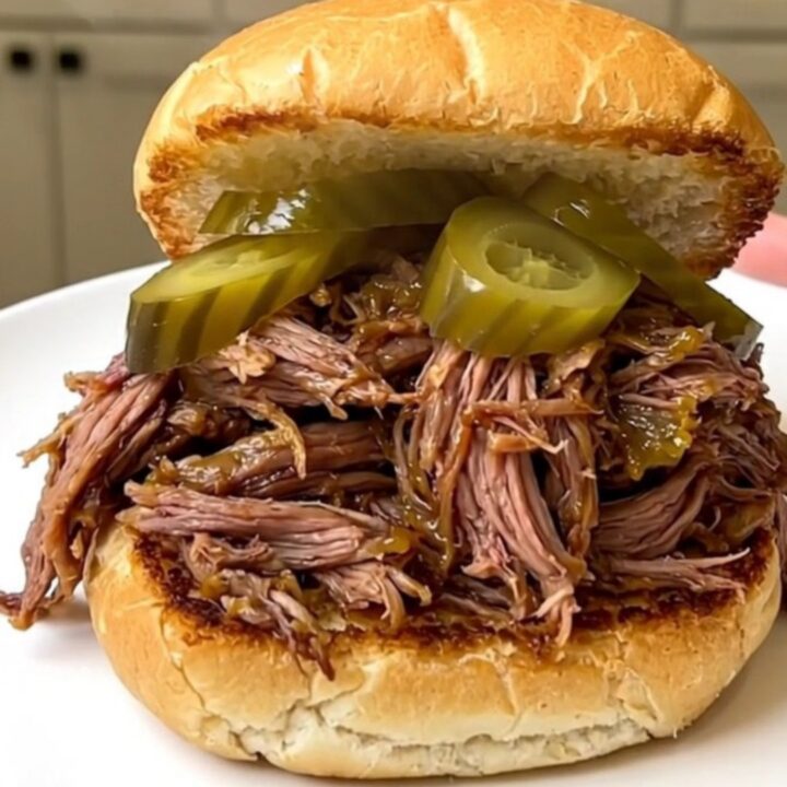 A close-up of a tender shredded beef sandwich on a toasted bun with pickle chips.