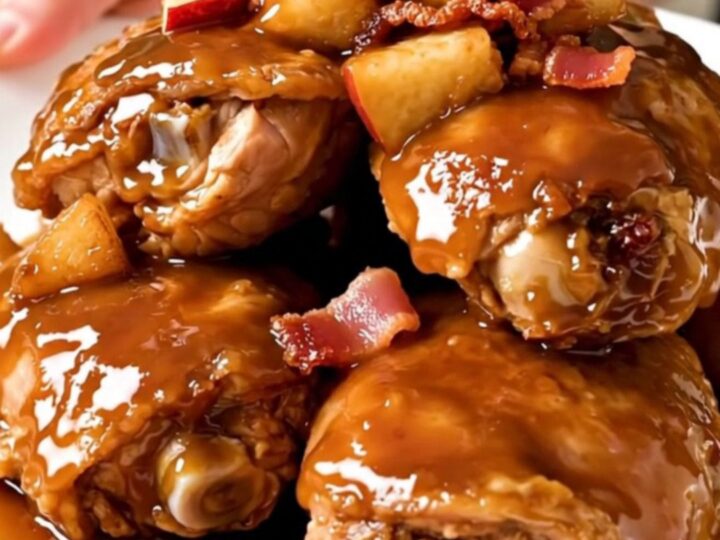A close-up shot of slow-cooked, glazed chicken thighs topped with tender apples and crispy bacon on a white plate.