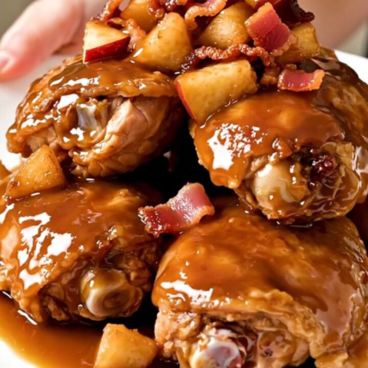 A close-up shot of slow-cooked, glazed chicken thighs topped with tender apples and crispy bacon on a white plate.