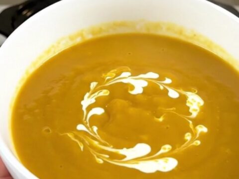 A white ceramic bowl filled with creamy orange pumpkin soup with a delicate swirl of heavy cream on top.