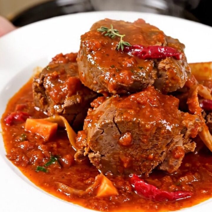 A close-up of a slow cooker filled with veal shanks, diced carrots, celery, and a rich tomato sauce.