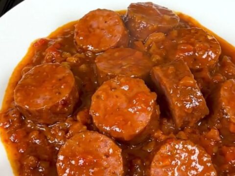 A top-down view of a slow cooker filled with sliced smoked sausage, onions, spices, and crushed tomato pulp.
