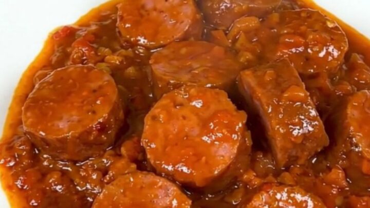 A top-down view of a slow cooker filled with sliced smoked sausage, onions, spices, and crushed tomato pulp.
