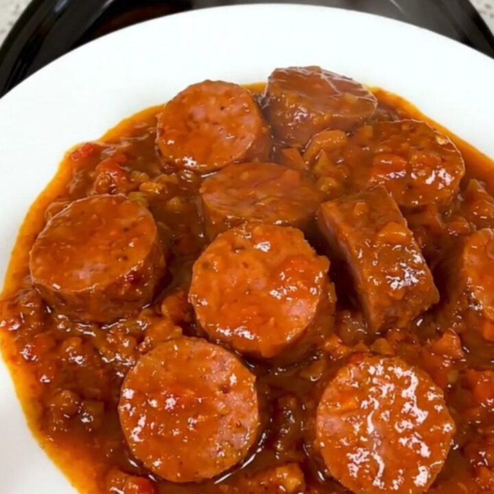 A top-down view of a slow cooker filled with sliced smoked sausage, onions, spices, and crushed tomato pulp.
