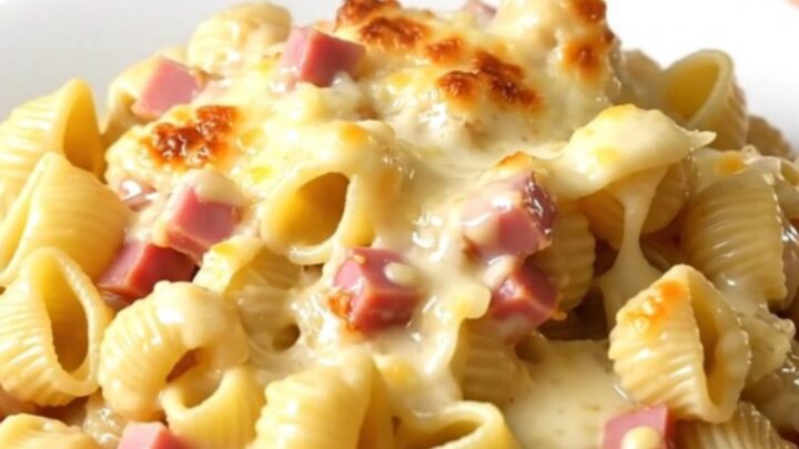 A top-down view of a black slow cooker filled with creamy pasta, ham, and melted cheese on a quartz countertop.