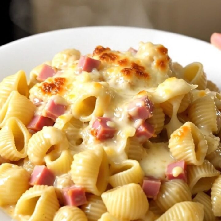 A top-down view of a black slow cooker filled with creamy pasta, ham, and melted cheese on a quartz countertop.