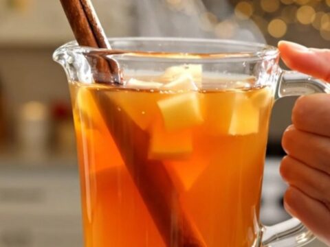 A clear glass mug filled with steaming hot apple cider, garnished with a cinnamon stick, sitting on a table.