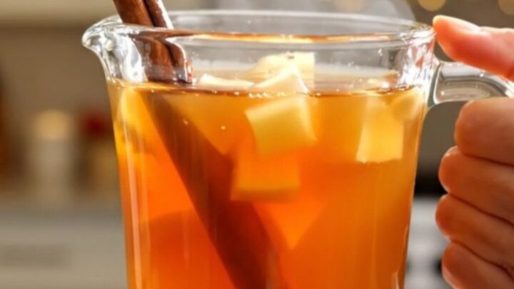 A clear glass mug filled with steaming hot apple cider, garnished with a cinnamon stick, sitting on a table.