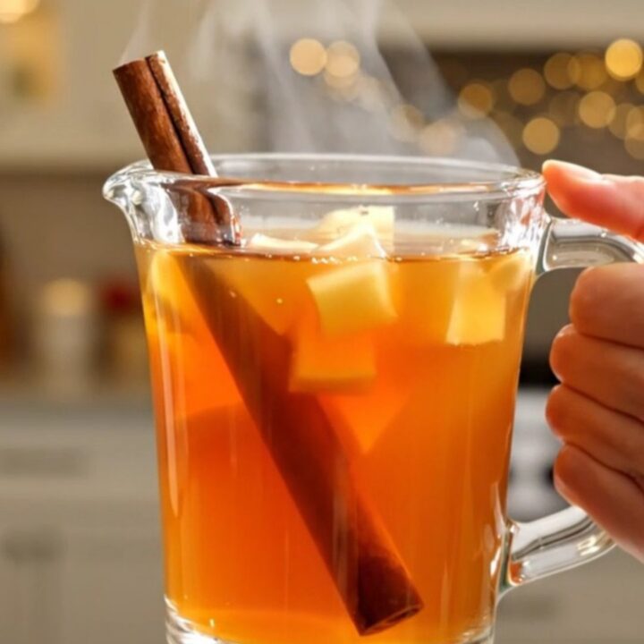 A clear glass mug filled with steaming hot apple cider, garnished with a cinnamon stick, sitting on a table.