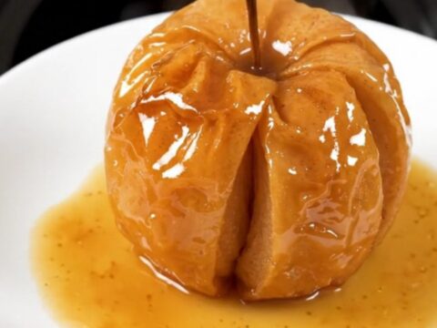 A close-up of a warm, glossy baked apple with cinnamon and brown sugar, served on a white plate.