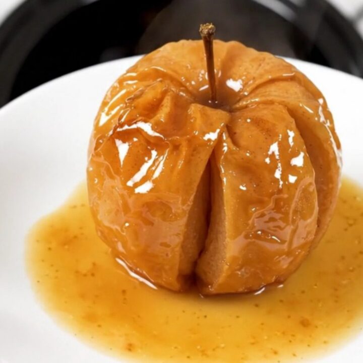 A close-up of a warm, glossy baked apple with cinnamon and brown sugar, served on a white plate.