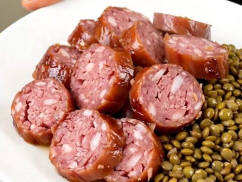 A top-down view of a slow cooker filled with Morteau sausages, green lentils, sliced carrots, and bay leaves.