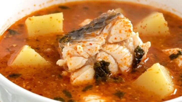 A top-down view of a black slow cooker filled with white fish, sliced potatoes, and a rich tomato broth.