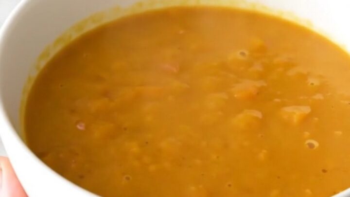 A bowl of creamy, bright orange red kuri squash and chestnut soup on a white table.