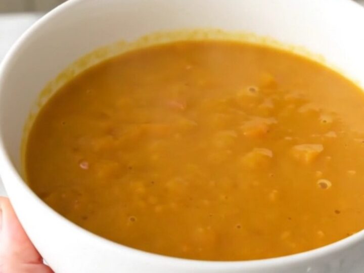 A bowl of creamy, bright orange red kuri squash and chestnut soup on a white table.