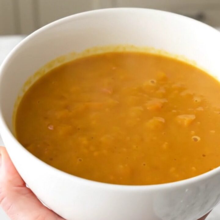 A bowl of creamy, bright orange red kuri squash and chestnut soup on a white table.