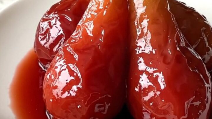 Four ruby-red poached pears sitting upright on a white plate with a glossy wine reduction syrup.