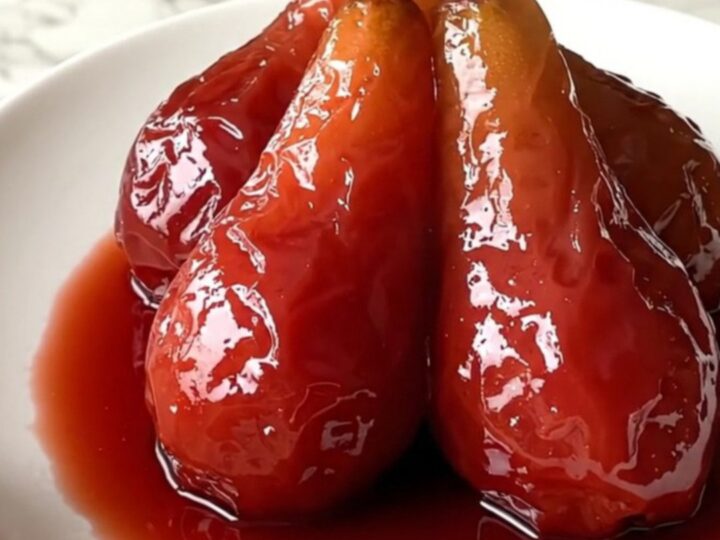 Four ruby-red poached pears sitting upright on a white plate with a glossy wine reduction syrup.