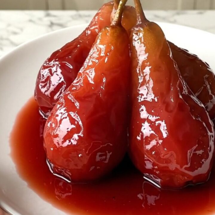 Four ruby-red poached pears sitting upright on a white plate with a glossy wine reduction syrup.