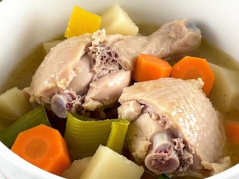A white ceramic bowl filled with tender chicken thighs, sliced carrots, leeks, and turnips in a savory broth.