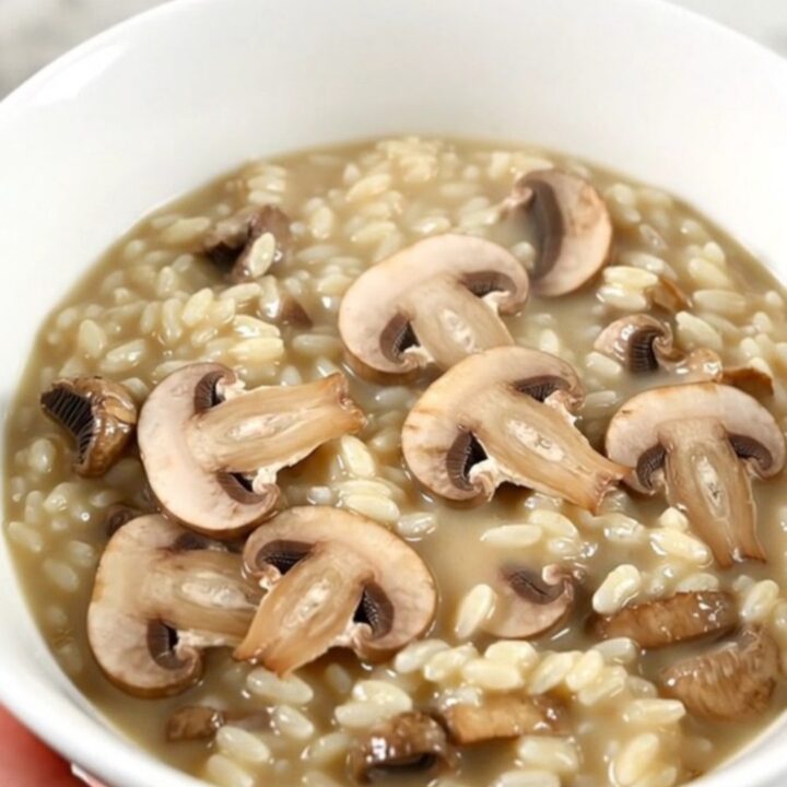 A top-down view of a black slow cooker filled with creamy mushroom risotto on a white speckled countertop.