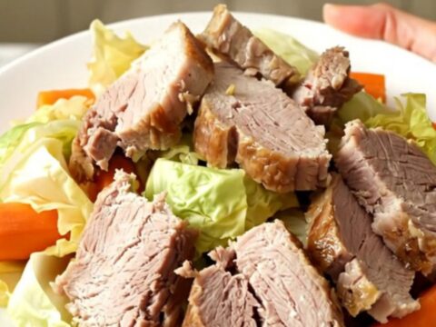 A slow cooker filled with tender salted pork, cabbage wedges, and halved carrots on a speckled quartz countertop.