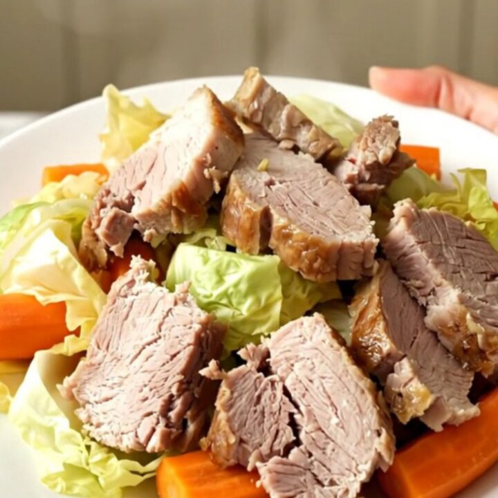 A slow cooker filled with tender salted pork, cabbage wedges, and halved carrots on a speckled quartz countertop.
