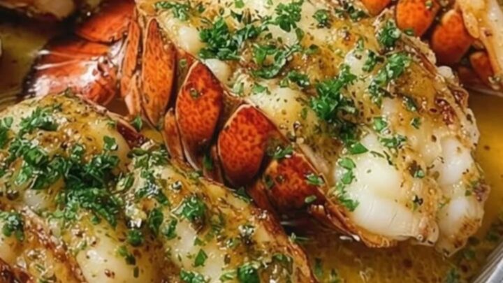 Four garlic herb baked lobster tails arranged in a foil pan, drizzled with golden butter and fresh parsley.