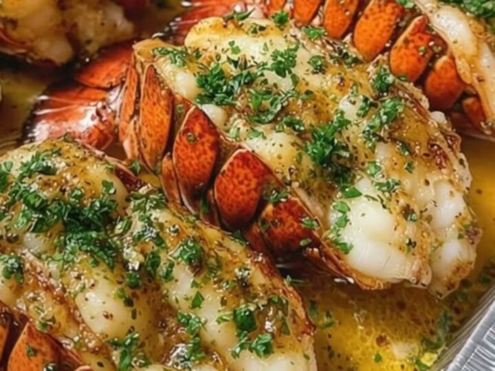 Four garlic herb baked lobster tails arranged in a foil pan, drizzled with golden butter and fresh parsley.