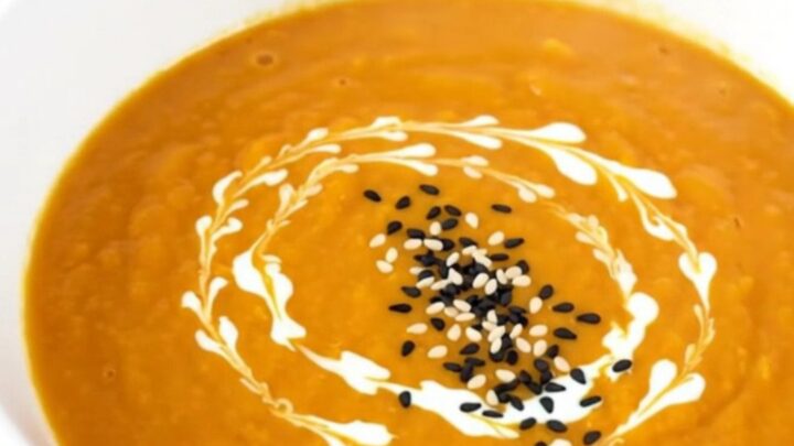 A white bowl filled with smooth, orange carrot soup garnished with a swirl of cream and toasted sesame seeds.