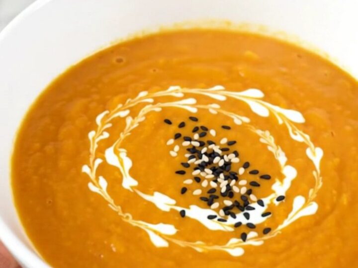 A white bowl filled with smooth, orange carrot soup garnished with a swirl of cream and toasted sesame seeds.