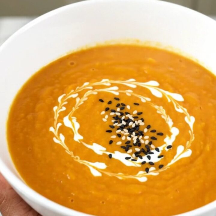A white bowl filled with smooth, orange carrot soup garnished with a swirl of cream and toasted sesame seeds.
