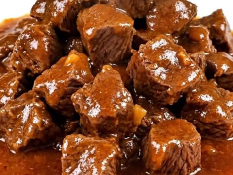 A steaming, glossy beef goulash with tender beef cubes and soft vegetables on a white plate.