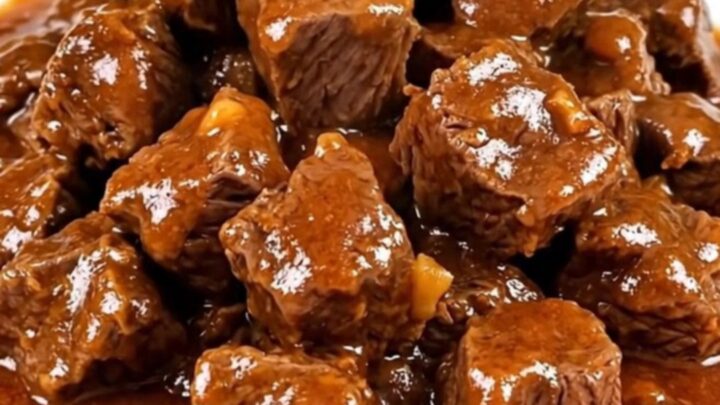 A steaming, glossy beef goulash with tender beef cubes and soft vegetables on a white plate.