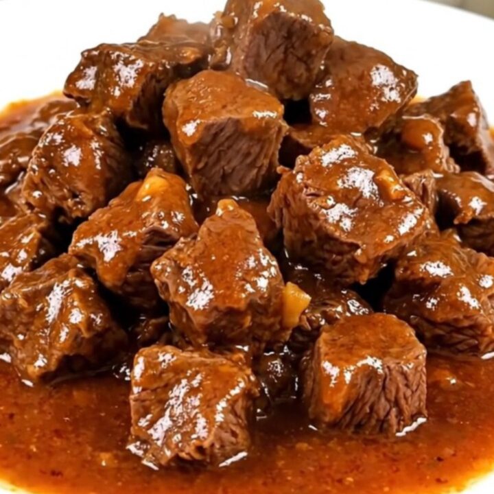 A steaming, glossy beef goulash with tender beef cubes and soft vegetables on a white plate.