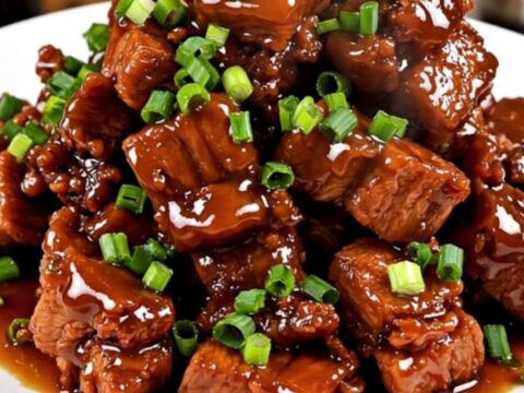 A close-up shot of glossy, caramelized pork cubes served on a white plate and garnished with fresh green scallions.