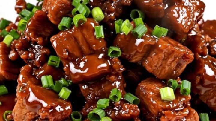 A close-up shot of glossy, caramelized pork cubes served on a white plate and garnished with fresh green scallions.