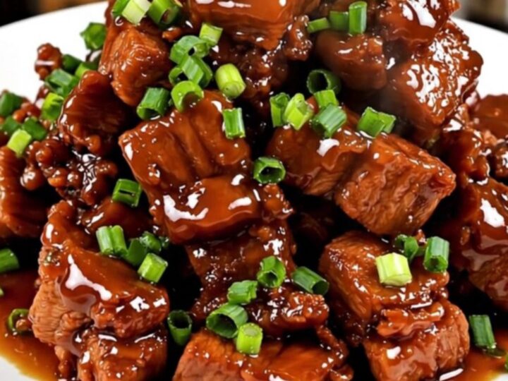A close-up shot of glossy, caramelized pork cubes served on a white plate and garnished with fresh green scallions.