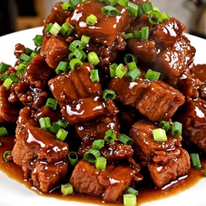 A close-up shot of glossy, caramelized pork cubes served on a white plate and garnished with fresh green scallions.