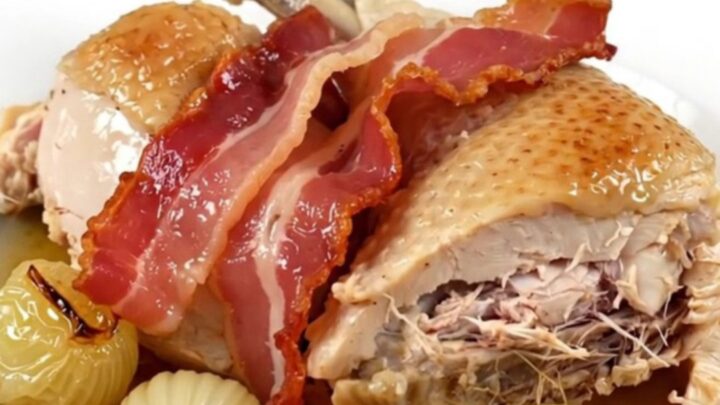 A top-down view of chicken thighs, bacon, and pearl onions cooking in a slow cooker.
