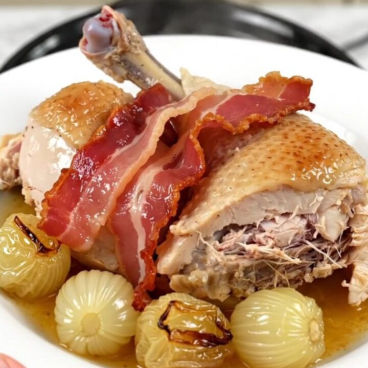 A top-down view of chicken thighs, bacon, and pearl onions cooking in a slow cooker.
