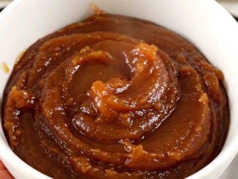 A white bowl filled with glossy, dark brown apple butter, steaming slightly in front of blurred kitchen cabinets.