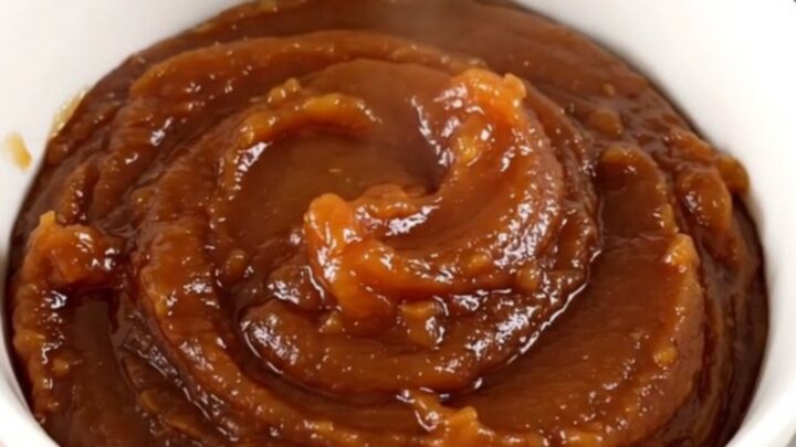 A white bowl filled with glossy, dark brown apple butter, steaming slightly in front of blurred kitchen cabinets.